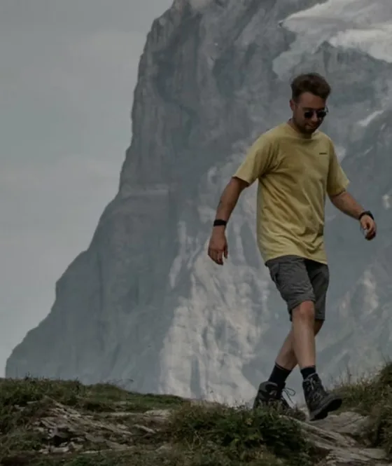 Randonneur en t-shirt jaune descendant une crête herbeuse face à une haute paroi rocheuse